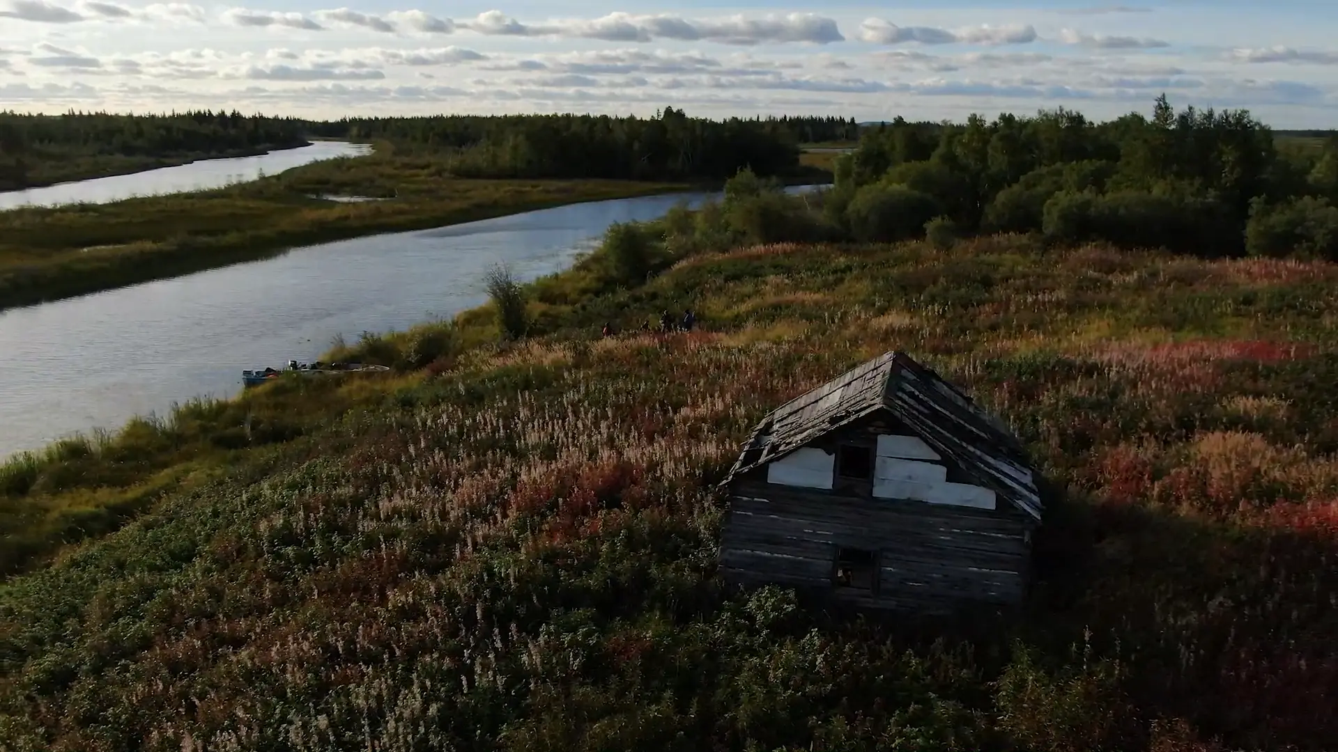 Image prise par un drone d'un ancien camp de traite situé sur la rive de la rivière Marian au coucher du soleil. La cabane en bois barricadée semble n'avoir été habitée par personne depuis des décennies. Une faune rouge, orange et jaune parsème la clairière, avec la rivière Marian visible en arrière-plan.