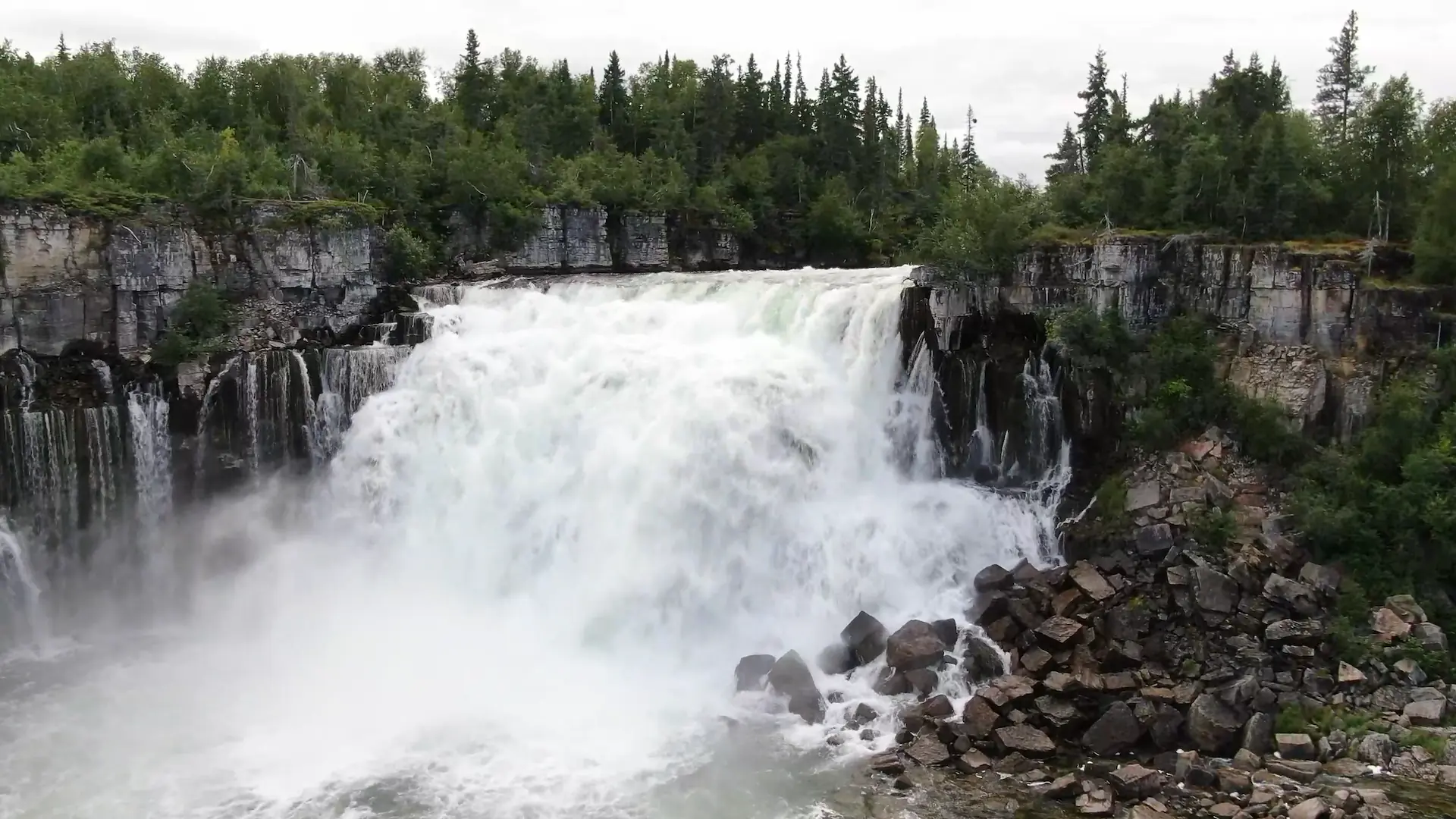 An aerial view of the Lac La Martre river plunging over one of the two 15 foot drops that make up Nàı̨lı̨ı̨ (Whatì Falls). Smaller streams of water pour out of the rocky walls on each side of the river.