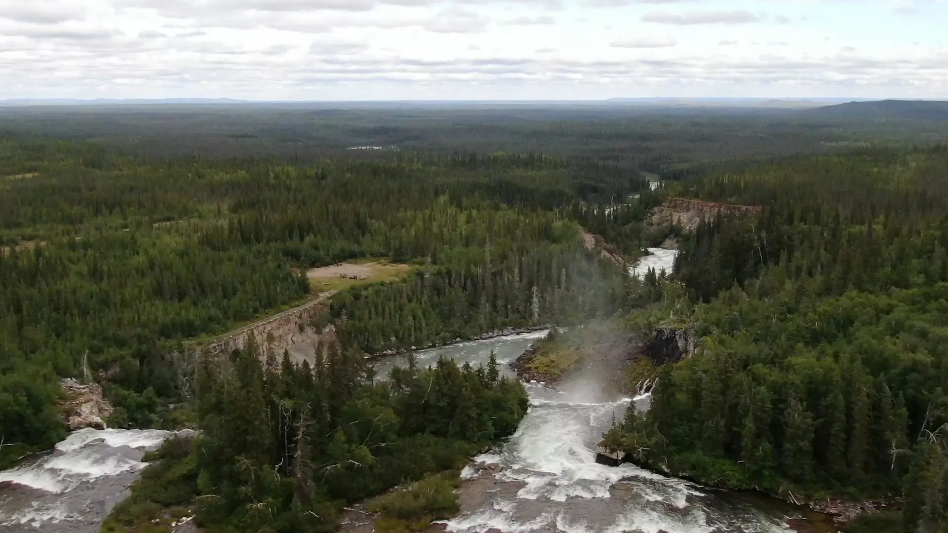 An overhead drone photography image of Whatì Falls. Mist rises from where the white water plunges over a cliff. Past the falls, a dirt parking lot connected to the Whatì Falls bypass road is visible. 