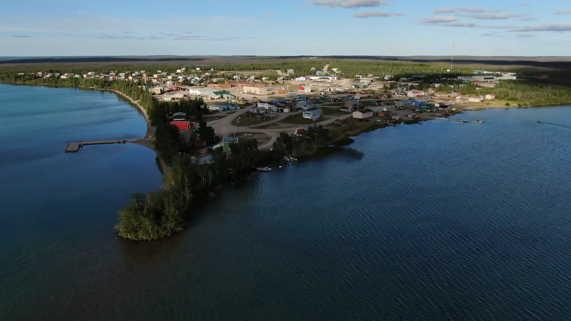 Une image de photographie de drone des eaux bleues claires du lac La Martre et de la communauté Tłı̨chǫ de Whatì. Des chemins de terre relient les maisons et les bâtiments communautaires de cette petite ville d'environ 500 habitants. Les bâtiments colorés parsèment la ville sans disposition particulière.