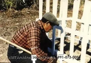 Vieille photo d'un homme en chemise à carreaux bruns et casquette grise s'agenouillant pour réparer la clôture blanche autour de la tombe.