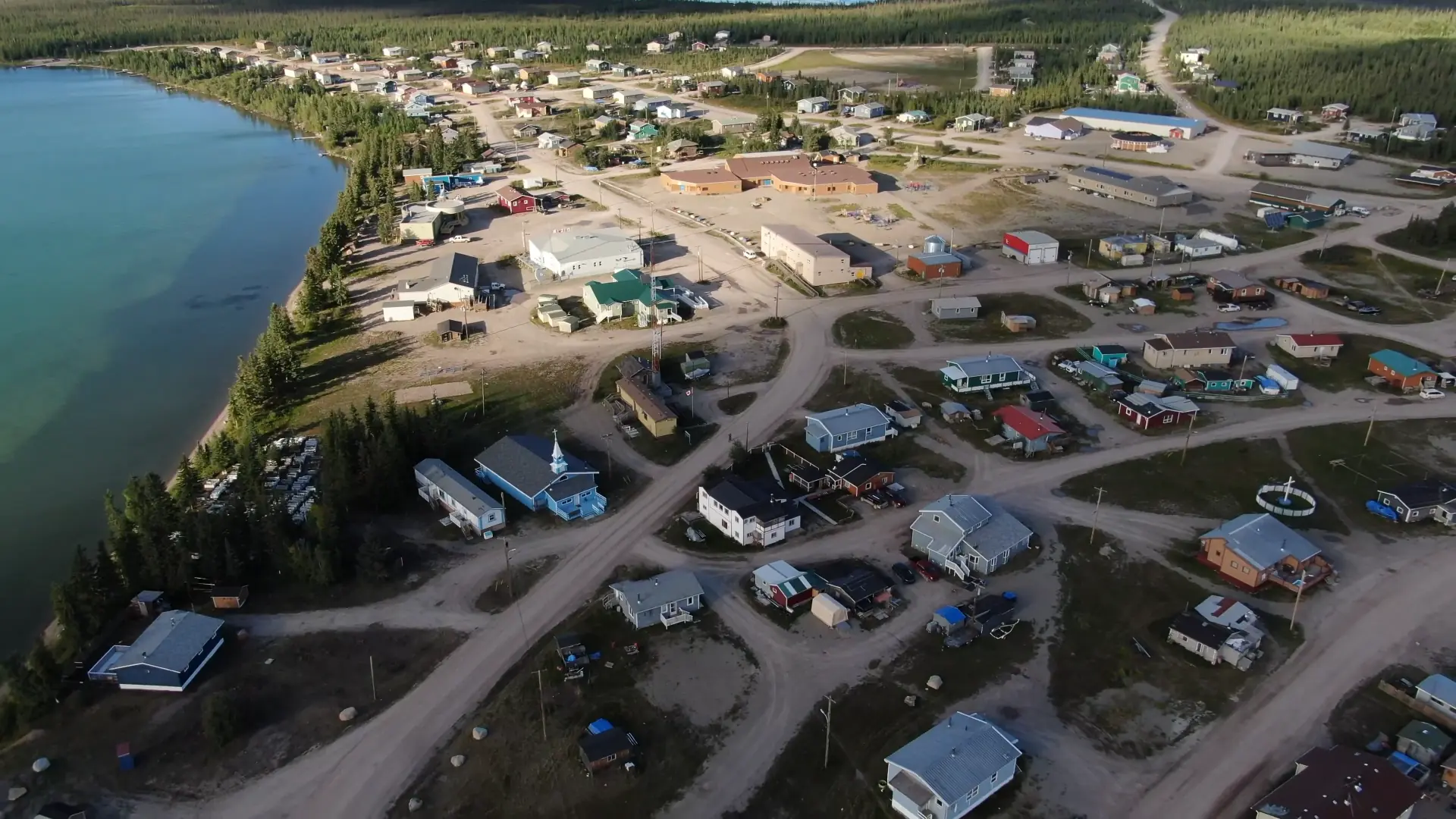 Aerial view of the community of Whatì showing dirt roads connecting houses and community buildings. The edge of the lake is visible on the left, lined with trees.